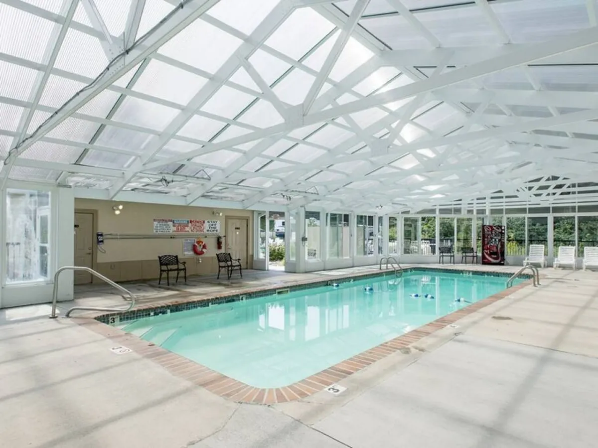 A bright and inviting indoor swimming pool features large windows that let in natural light, white wooden beams overhead, and several lounge chairs along the pool's edge. The tranquil atmosphere is perfect for year-round swimming and socializing with family and friends.