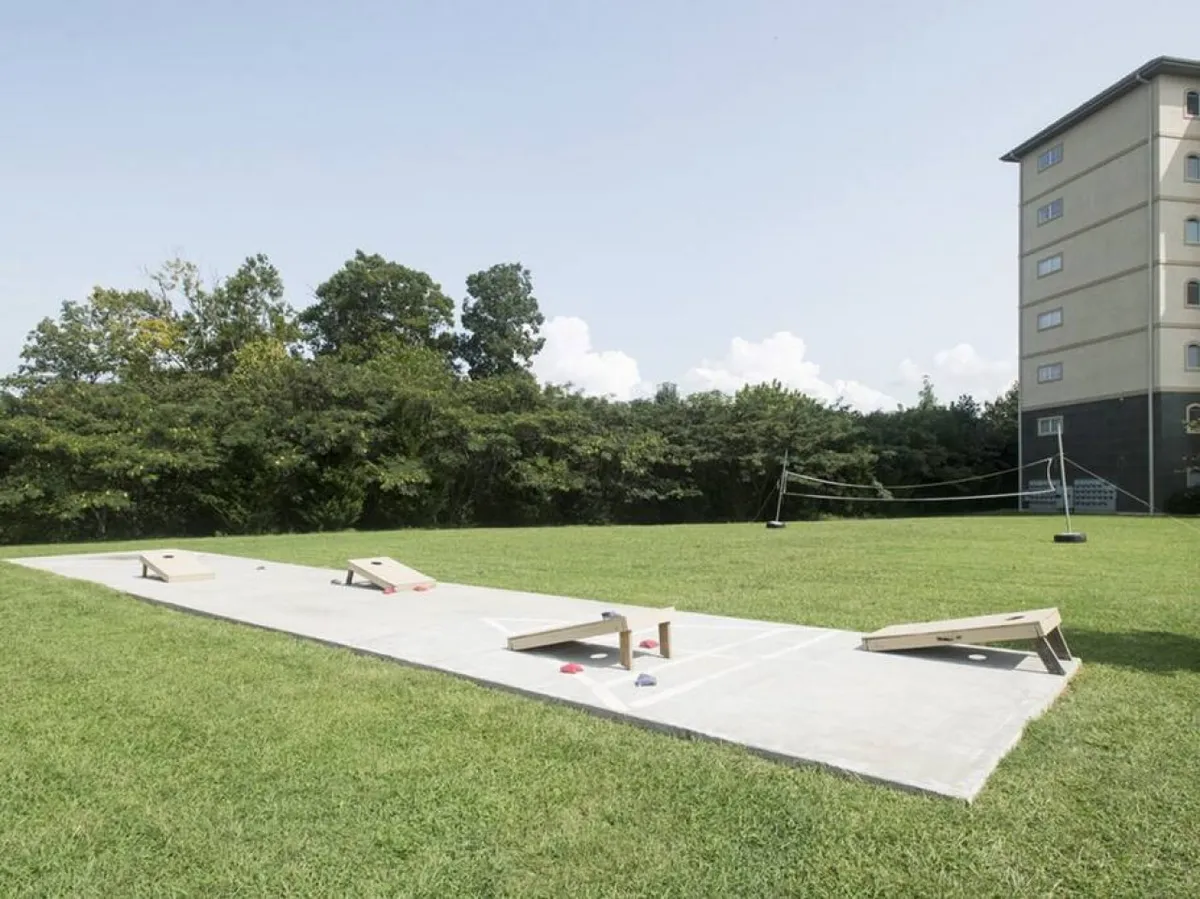 An active outdoor recreation area featuring cornhole boards, a grassy space, and a volleyball net, with the condo building in the background. The scene captures outdoor play and social gatherings.