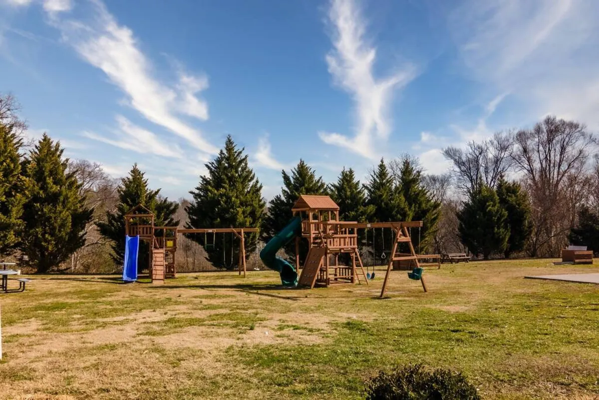 A cheerful playground with a slide, swings, and tall trees providing shade. The inviting green grass and open layout offer plenty of space for children to play, while families gather nearby for picnics and outdoor games, emphasizing a joyful, community-oriented atmosphere.