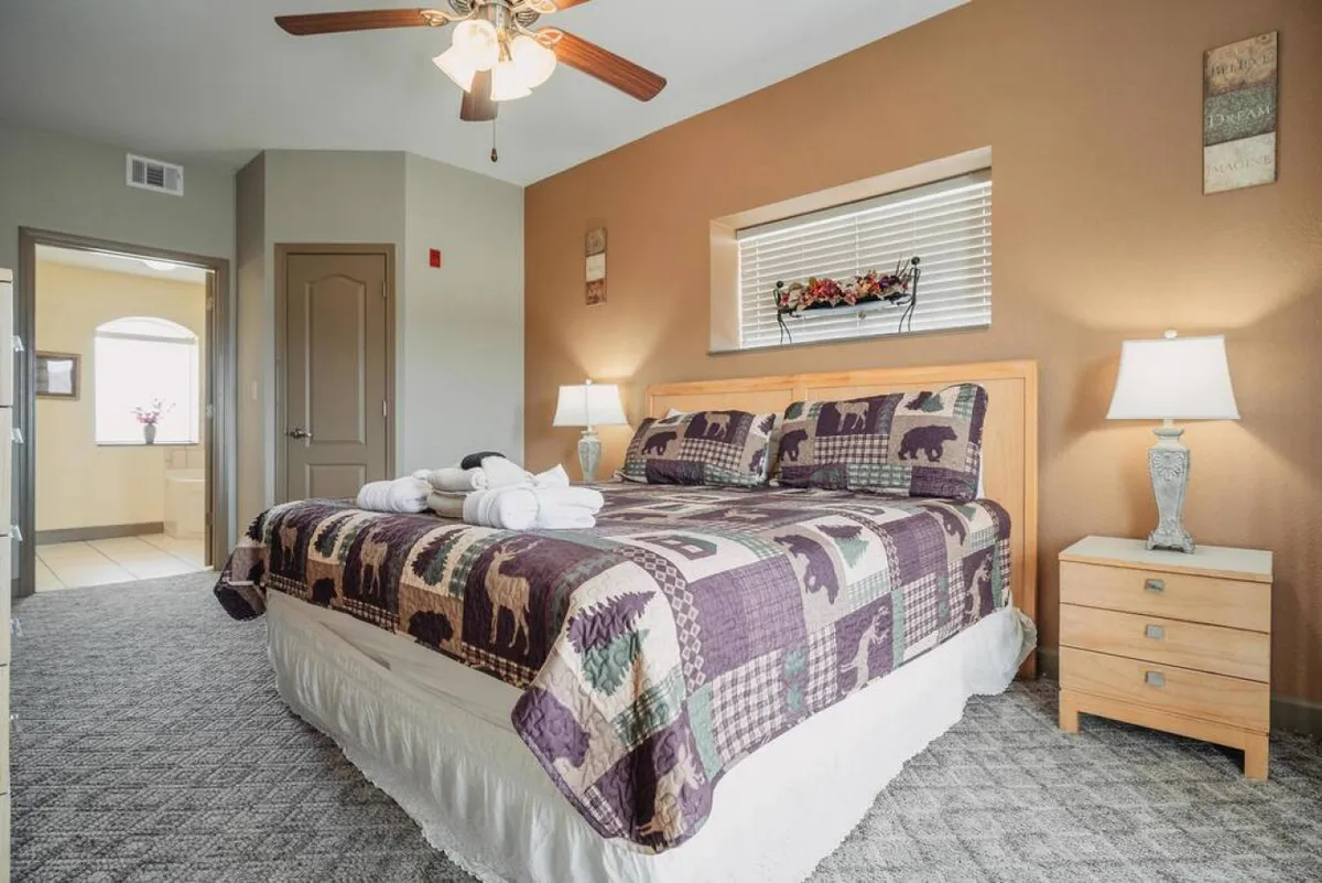 A welcoming bedroom accented with a bed adorned in a patterned quilt, flanked by nightstands with lamps and decorative wall accents. Soft natural light streams in, enhancing the comforting and restful mood of the space.