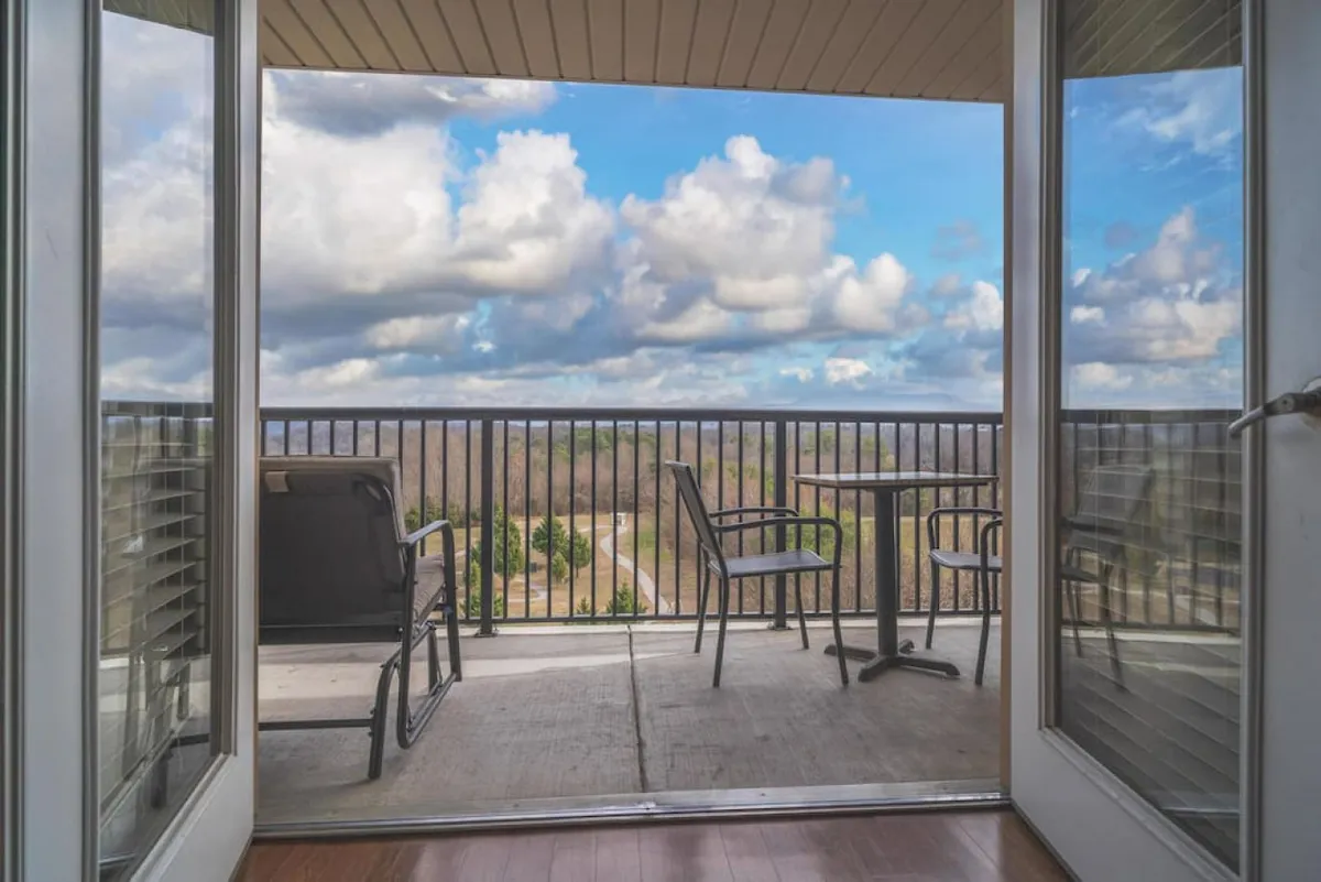 The image showcases the condo's private balcony with a railing, a table and chairs set up for guests, and an expansive view of the lush mountain landscape beneath a cloudy sky. The scene suggests a tranquil spot for relaxing outdoors, sipping coffee, and soaking in natural beauty.