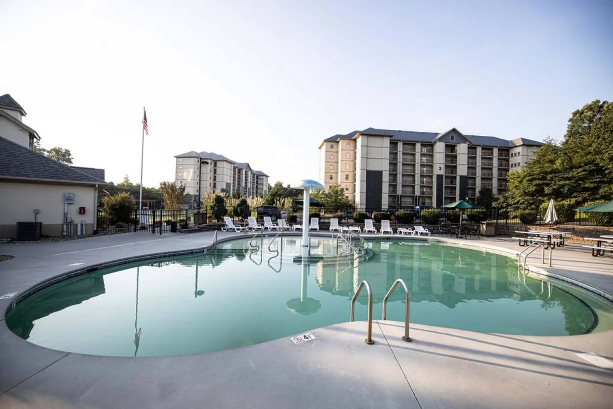 A vibrant view of the resort-style swimming pool area, complete with lounge chairs and lush greenery. Buildings are visible in the background, suggesting proximity to the main accommodations.
