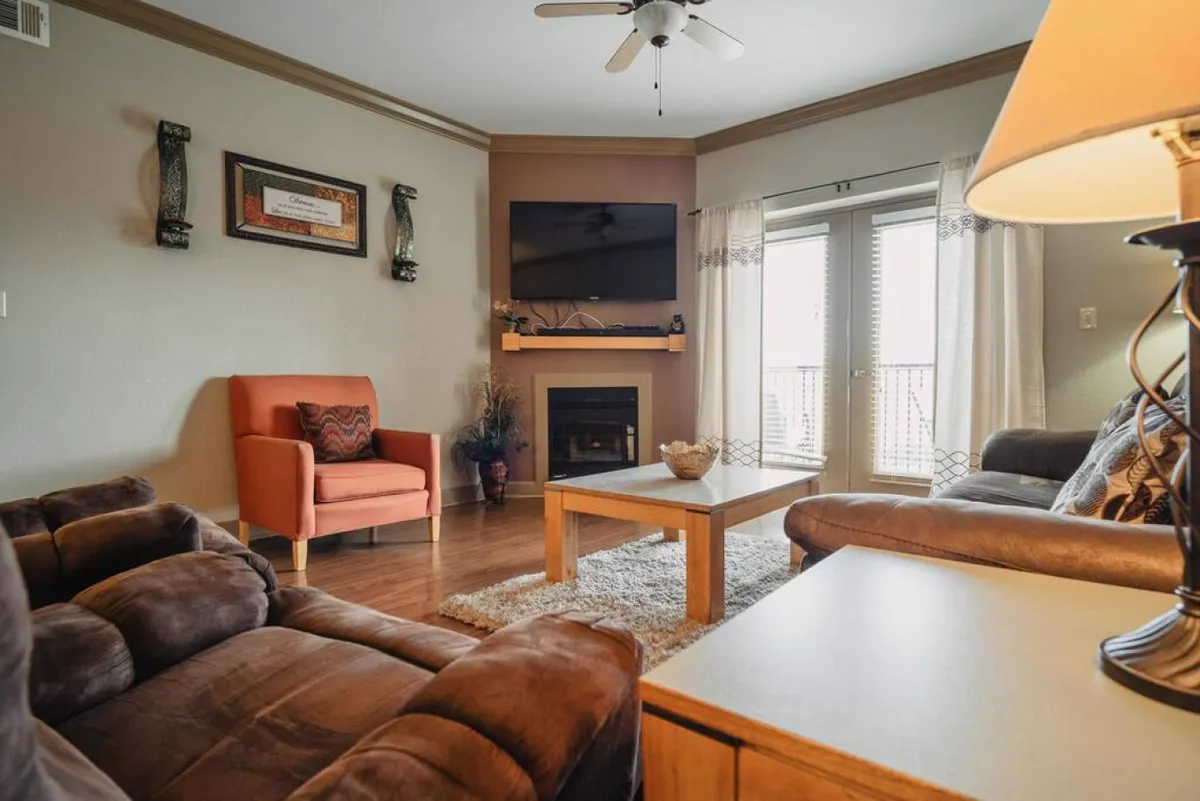 This image shows a comfortable seating area with a fireplace, television, and balcony doors that let in natural light. The setup is open and inviting, with ample space for movement and relaxation, suggesting a layout that could accommodate guests with diverse mobility needs.