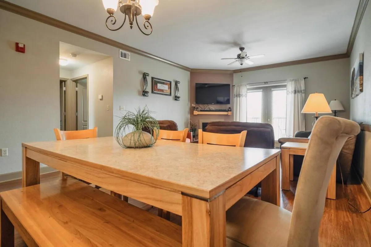 A spacious dining area with a wooden table and comfortable chairs, a ceiling fan overhead, and a large window letting in natural light. The living area is visible in the background, reinforcing the open, communal nature of the condo's interiors.