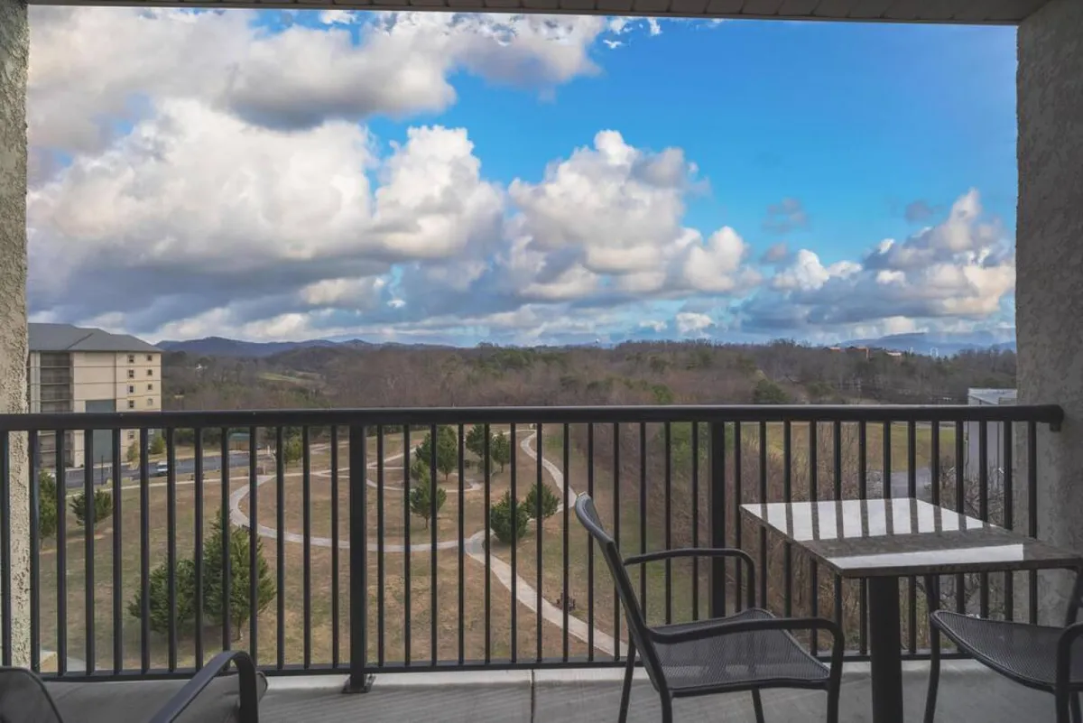 This image features a balcony with a table and chairs overlooking distant mountains and a cloud-filled sky, with a winding path below. The outdoor setting evokes both relaxation and the excitement of being near attractions, while the visible path subtly hints at ease of travel and exploration.