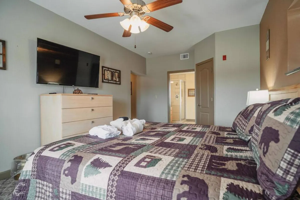 A spacious bedroom featuring a bed with a patterned quilt, a large TV mounted on the wall, and a dressing chest, all set in a cozy, homely atmosphere. The room exudes comfort, offering plenty of space for relaxation and entertainment.