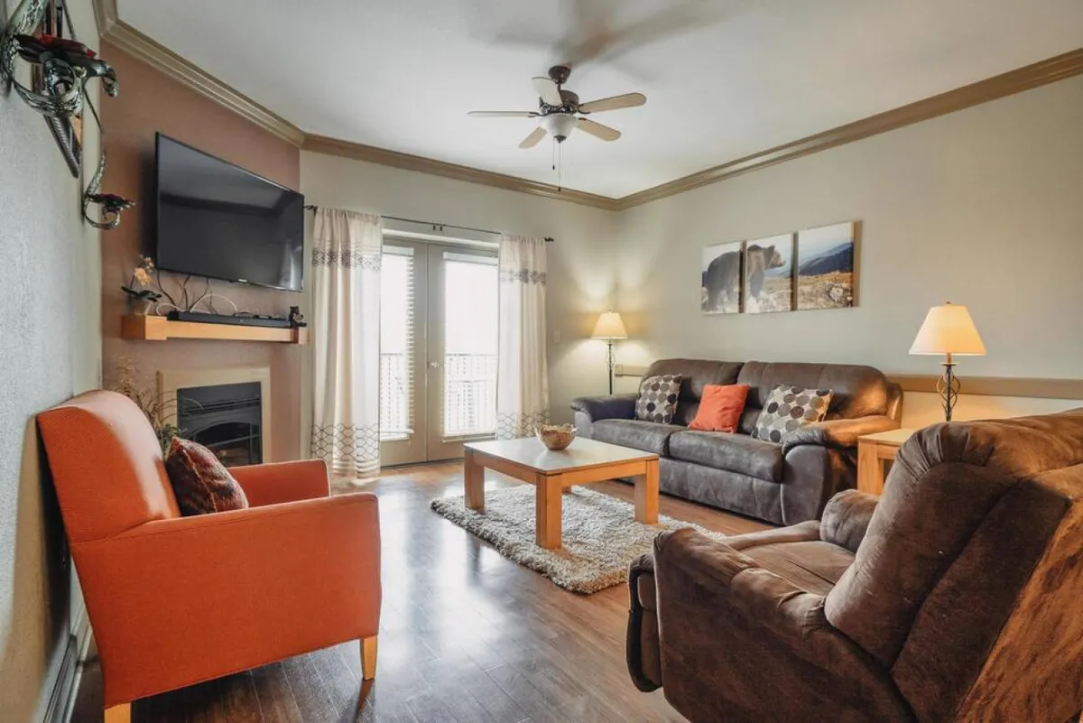 A cozy living space featuring a sofa, armchair, coffee table, television, fireplace, and decorative artwork. The room is filled with natural light from large windows, and the arrangement emphasizes easy flow and comfort, making it suitable for various guests, including those with mobility aids.