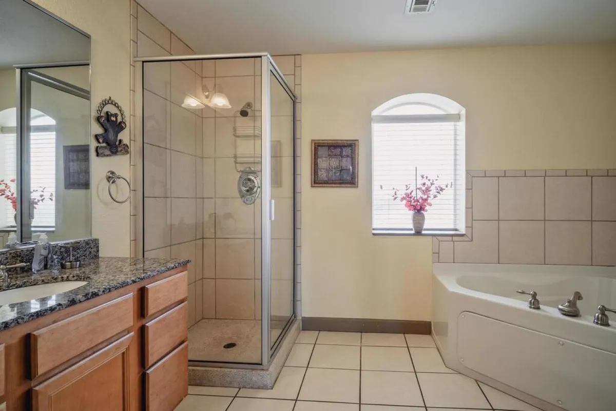 A beautifully appointed bathroom featuring a glass shower enclosure, a bathtub perfect for soaking, a granite countertop with a sink, decorative artwork, and a window with a floral arrangement. The space is tranquil and inviting, ideal for rejuvenation.